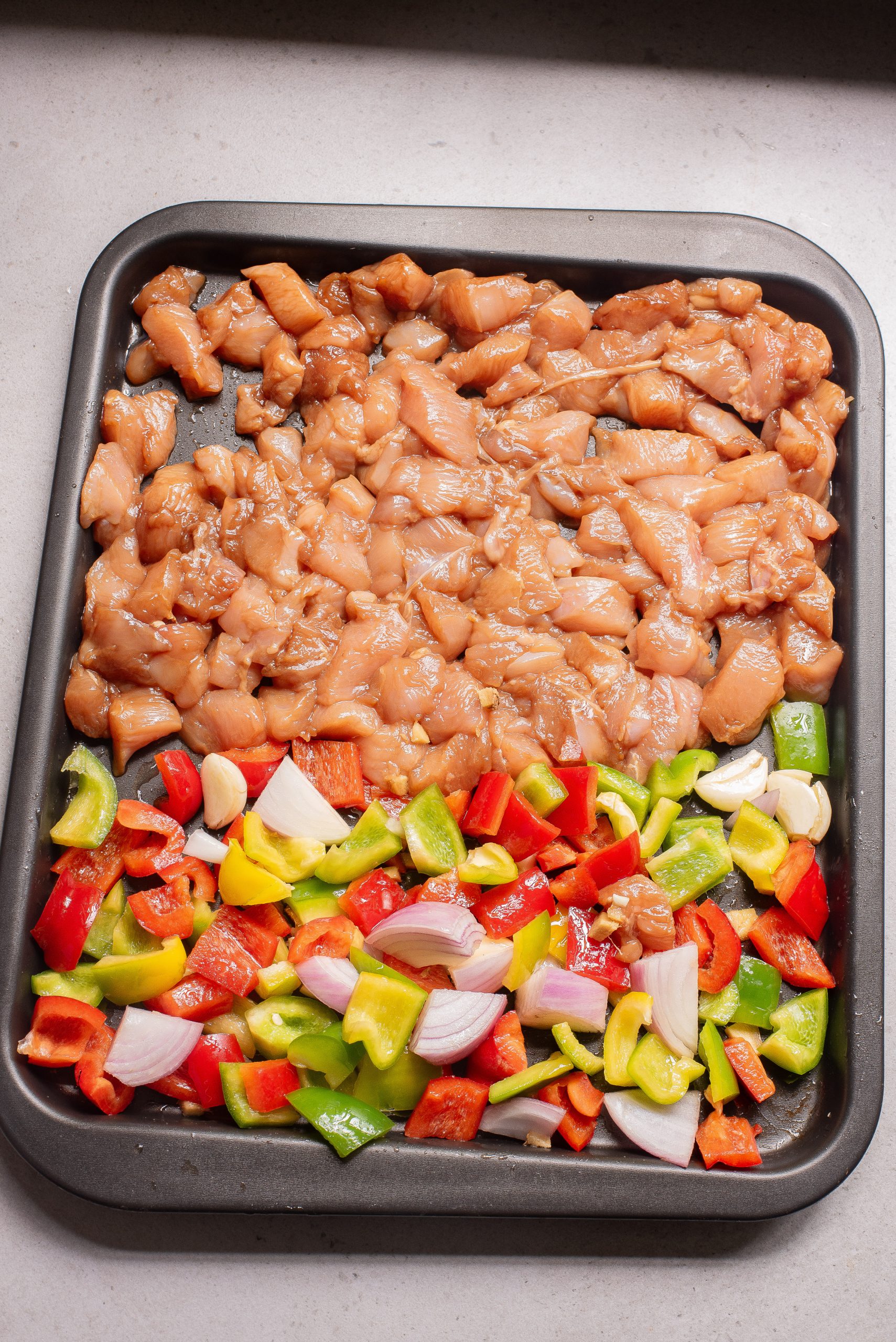 A baking tray with diced raw chicken on one side and chopped red, green, and yellow bell peppers and onions on the other—perfect for making Sheet Pan Bell Pepper Chicken.