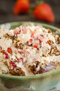 A close-up of a strawberry praline crunch salad, showcasing creamy dessert topped with chopped strawberries and crumbled brown pieces in a green bowl, with blurred strawberries in the background.
