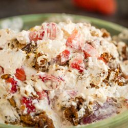 A close-up of a strawberry praline crunch salad, showcasing creamy dessert topped with chopped strawberries and crumbled brown pieces in a green bowl, with blurred strawberries in the background.