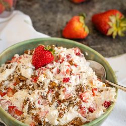 A bowl of creamy strawberry praline crunch salad mixed with chopped strawberries and nuts, topped with a whole strawberry, sits on a table with loose strawberries nearby.