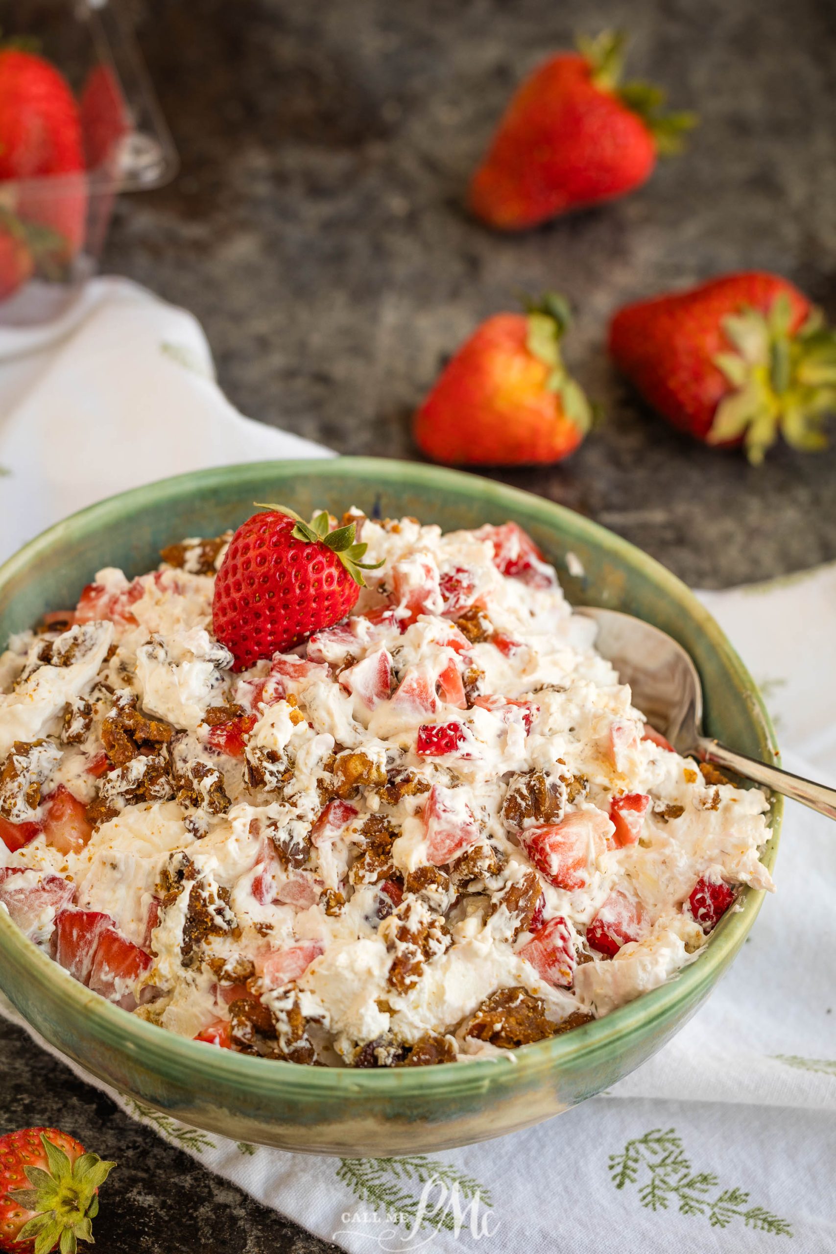 A bowl of fluff mixed with chopped strawberries and nuts, topped with a whole strawberry, sits on a table with loose strawberries nearby.