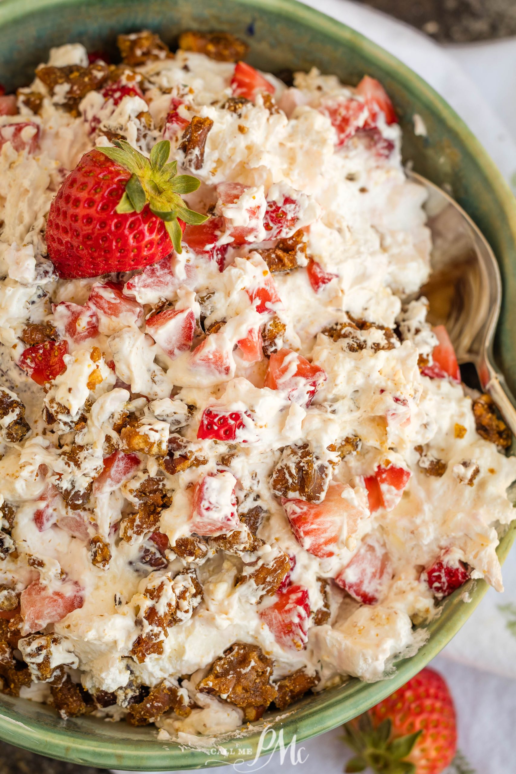 A bowl filled with a creamy strawberry praline crunch salad, mixed with chopped strawberries, crushed cookies or cereal, and topped with a whole strawberry. A spoon rests at the side of the bowl.