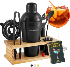 A black cocktail shaker set with tools in a wooden holder, a glass of orange cocktail with fruit, and a deck of cocktail recipe cards—perfect for those who love to make cocktails at home.