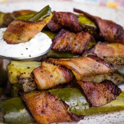 Bacon-wrapped jalapeño poppers and crispy bacon wrapped pickle spears on a plate with a small cup of white dipping sauce.