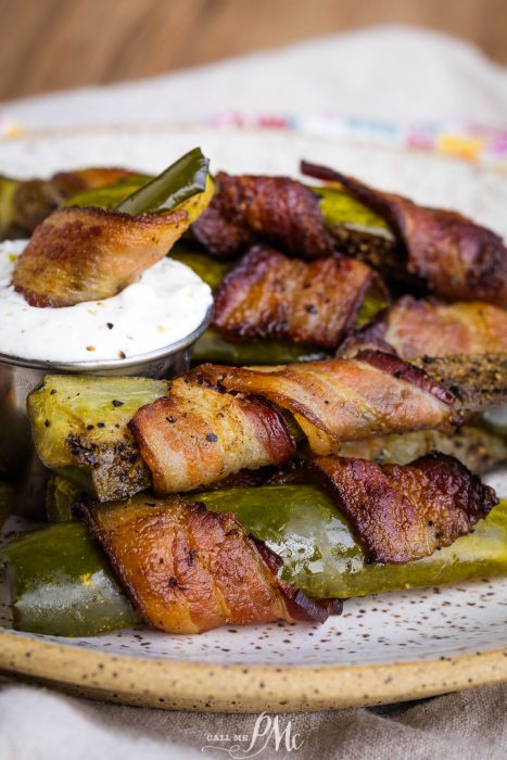 Bacon-wrapped jalapeño poppers and crispy bacon wrapped pickle spears on a plate with a small cup of white dipping sauce.