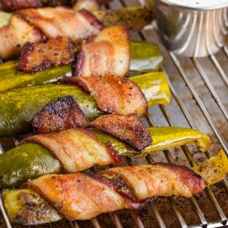 Jalapeño peppers and bacon wrapped pickle spears on a wire rack, with a cup of white dipping sauce topped with black pepper in the background.