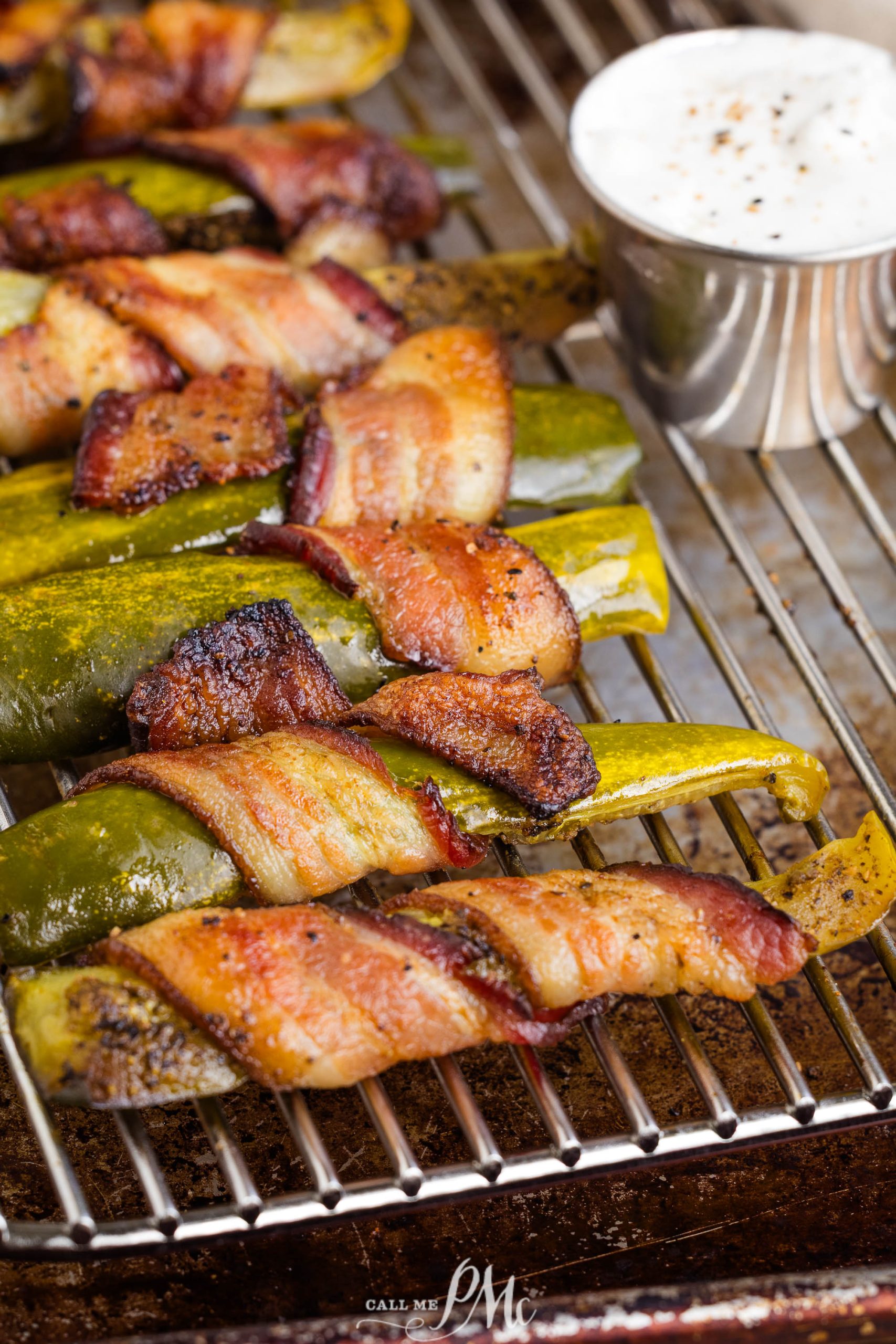 Jalapeño peppers and bacon wrapped pickle spears on a wire rack, with a cup of white dipping sauce topped with black pepper in the background.