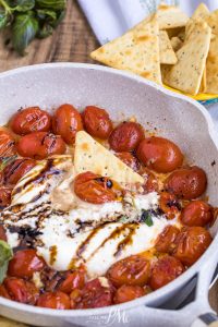 A skillet of Balsamic Tomato Burrata Dip features roasted cherry tomatoes, melted cheese, balsamic glaze, and a pita chip, with a bowl of pita chips in the background.