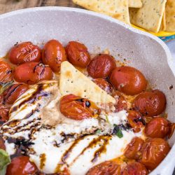 A skillet of Balsamic Tomato Burrata Dip features roasted cherry tomatoes, melted cheese, balsamic glaze, and a pita chip, with a bowl of pita chips in the background.
