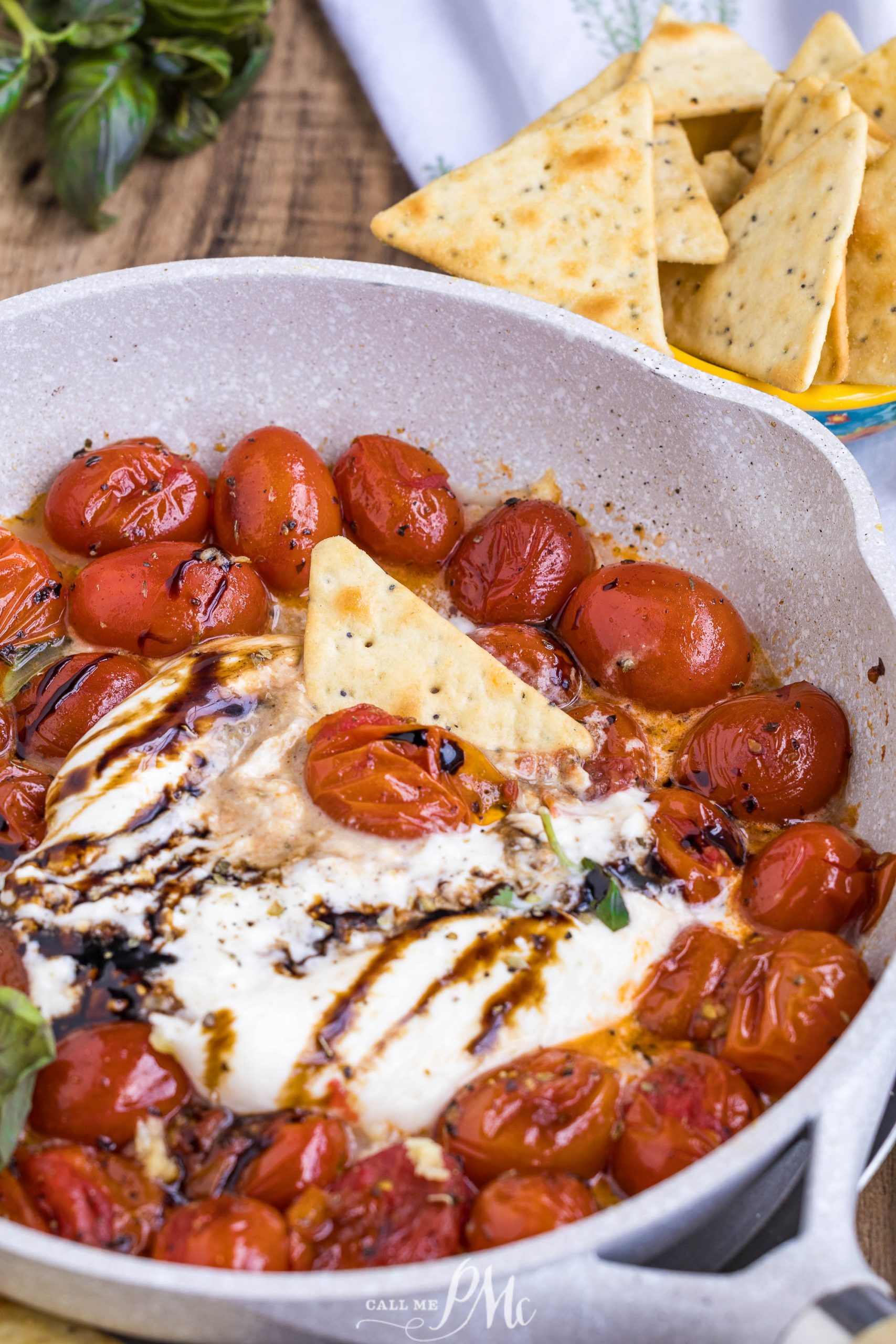 A skillet of Balsamic Tomato Burrata Dip features roasted cherry tomatoes, melted cheese, balsamic glaze, and a pita chip, with a bowl of pita chips in the background.