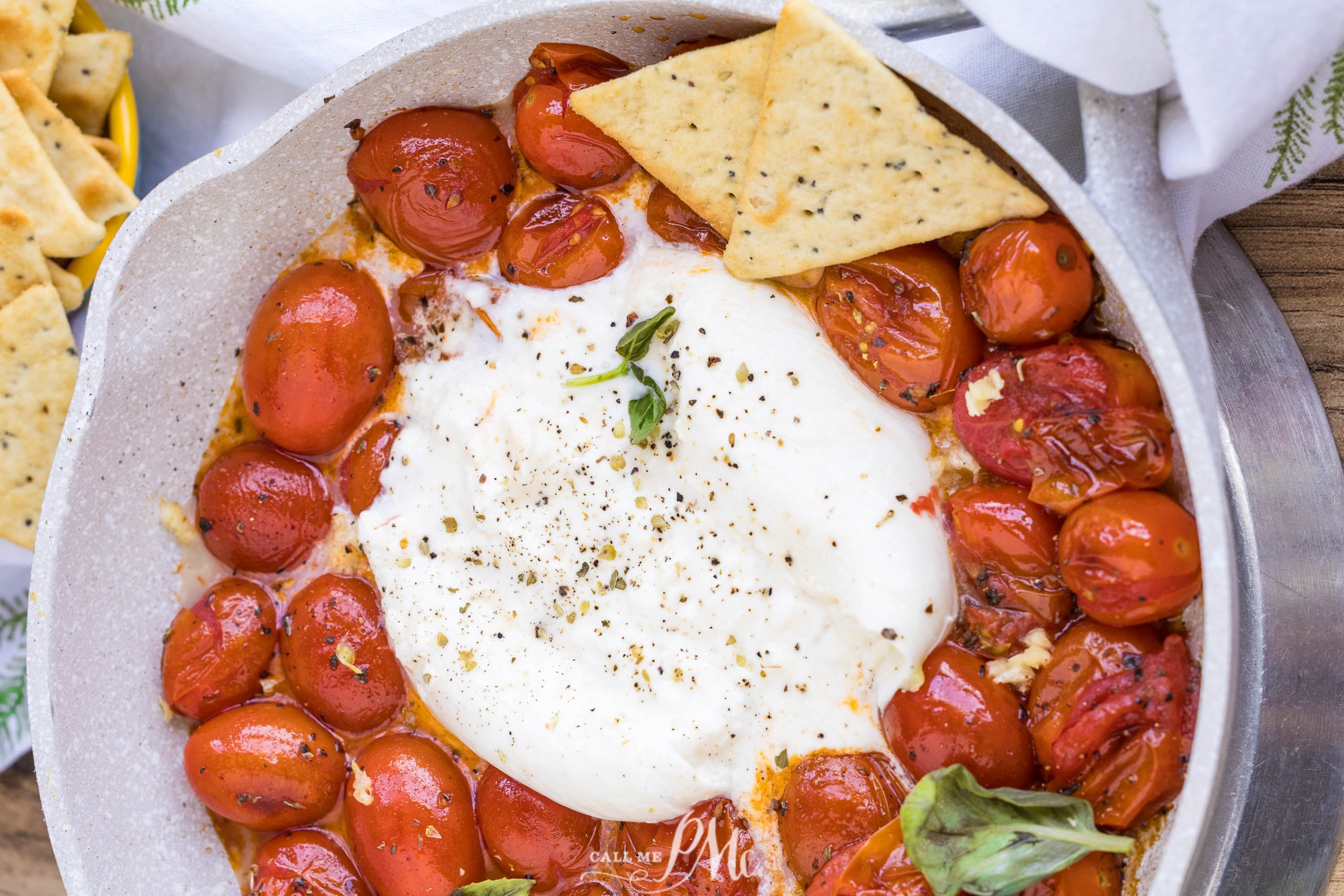A skillet filled with roasted cherry tomatoes and a large ball of burrata cheese in the center, garnished with herbs, makes this Balsamic Tomato Burrata Dip perfect for serving alongside crispy crackers.