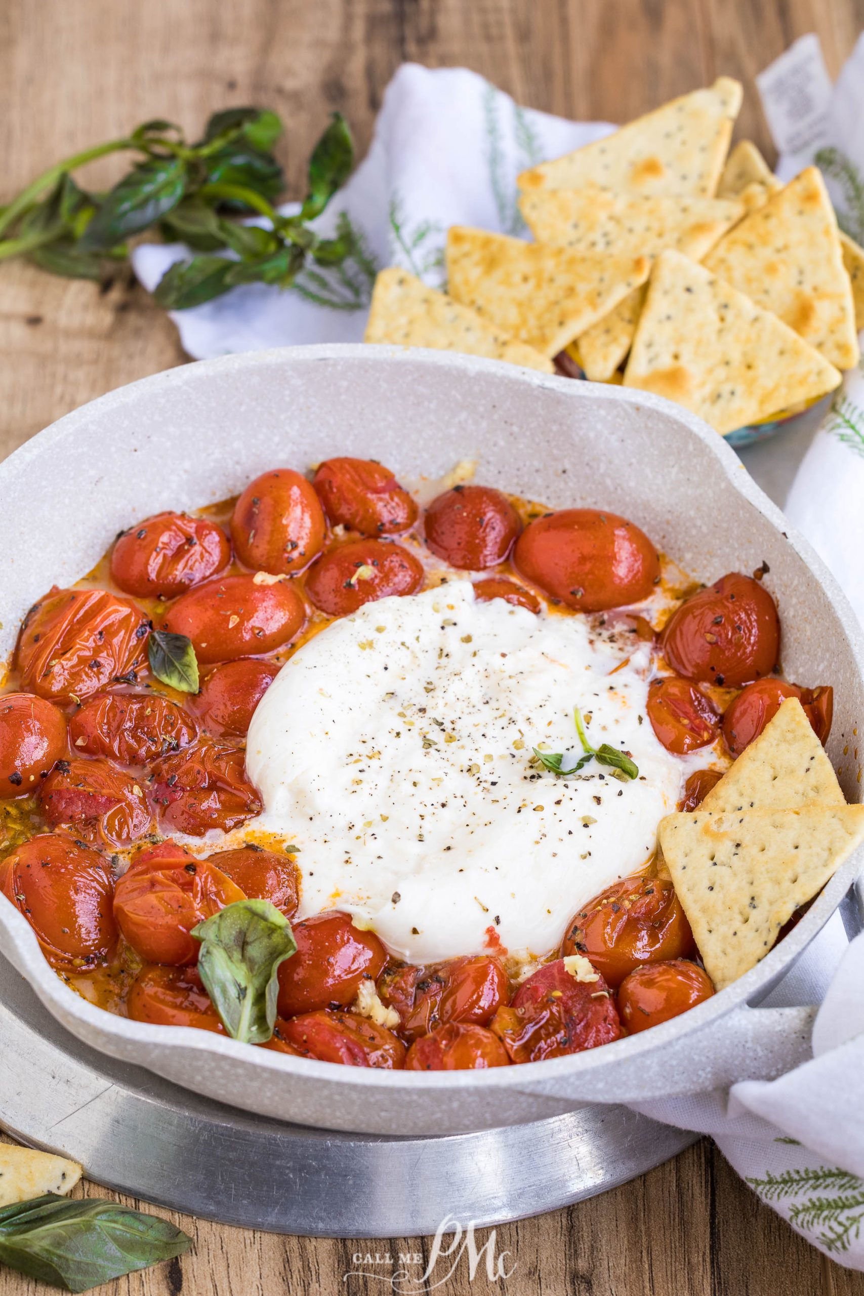A white frying pan filled with Balsamic Tomato Burrata Dip—roasted cherry tomatoes and a ball of burrata cheese, garnished with fresh basil, served with crackers on the side.
