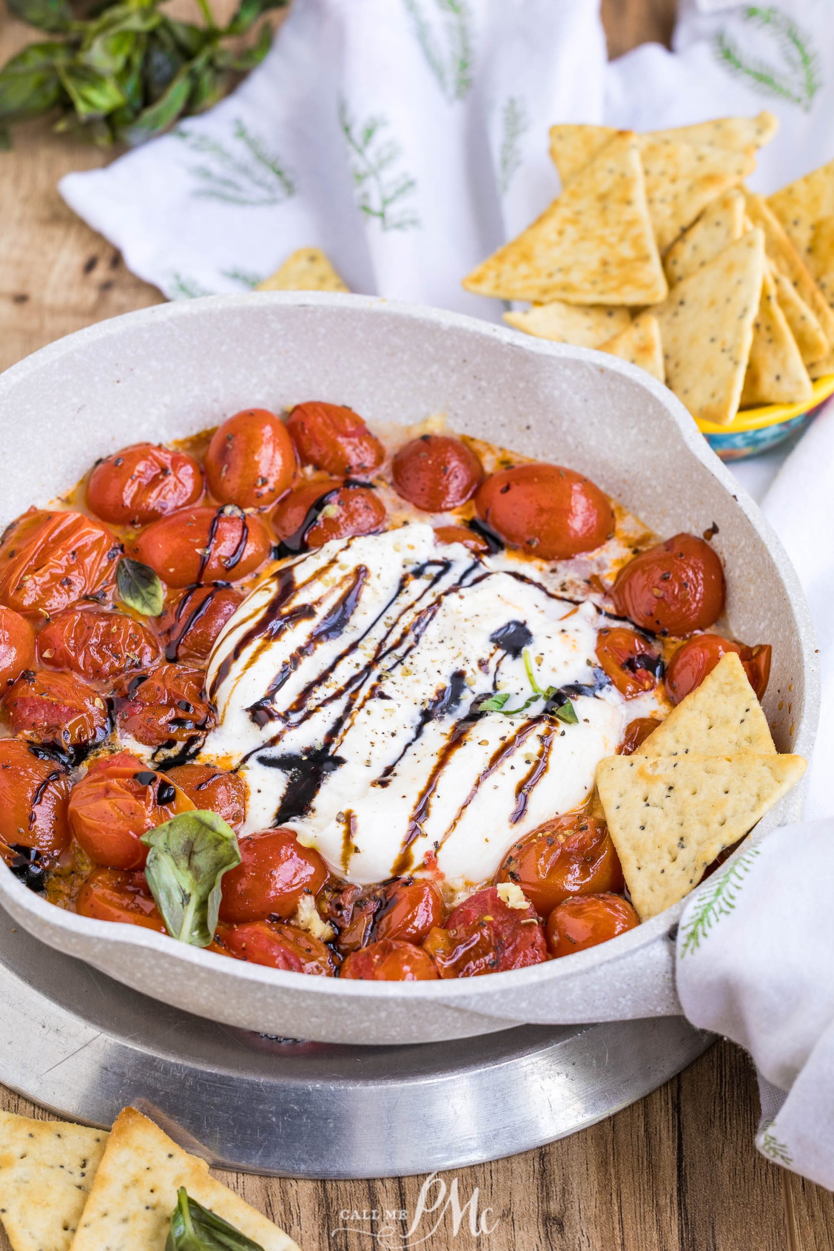 A skillet vegetables and cheese drizzled with balsamic glaze, fresh basil, and several rectangular crackers on the side.