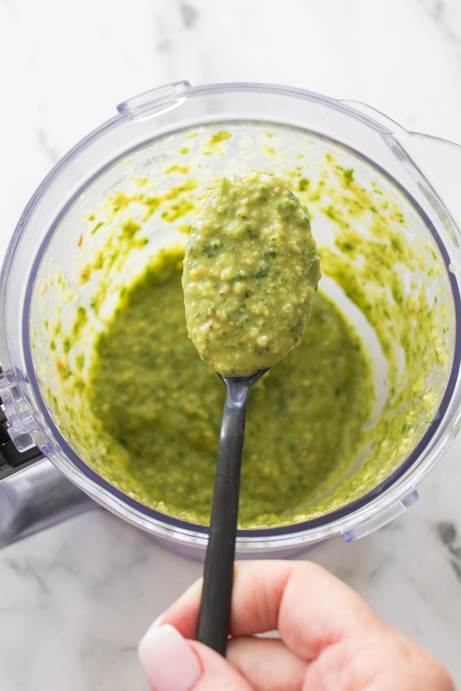 A hand holding a spoonful of thick green classic basil pesto over a food processor bowl filled with the same vibrant mixture.