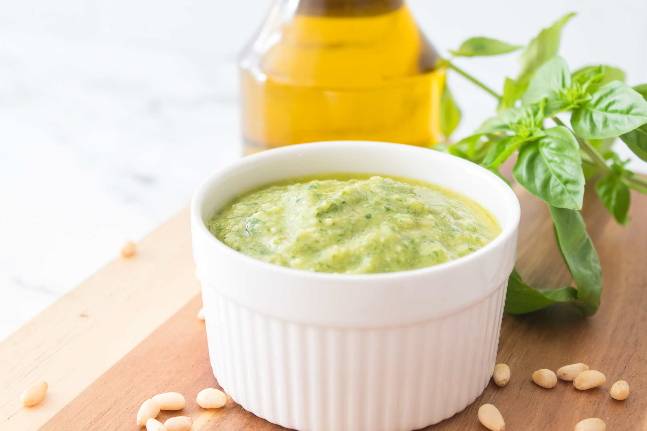 A white ramekin filled with classic basil pesto sauce sits on a wooden board next to fresh basil, pine nuts, and a bottle of oil.