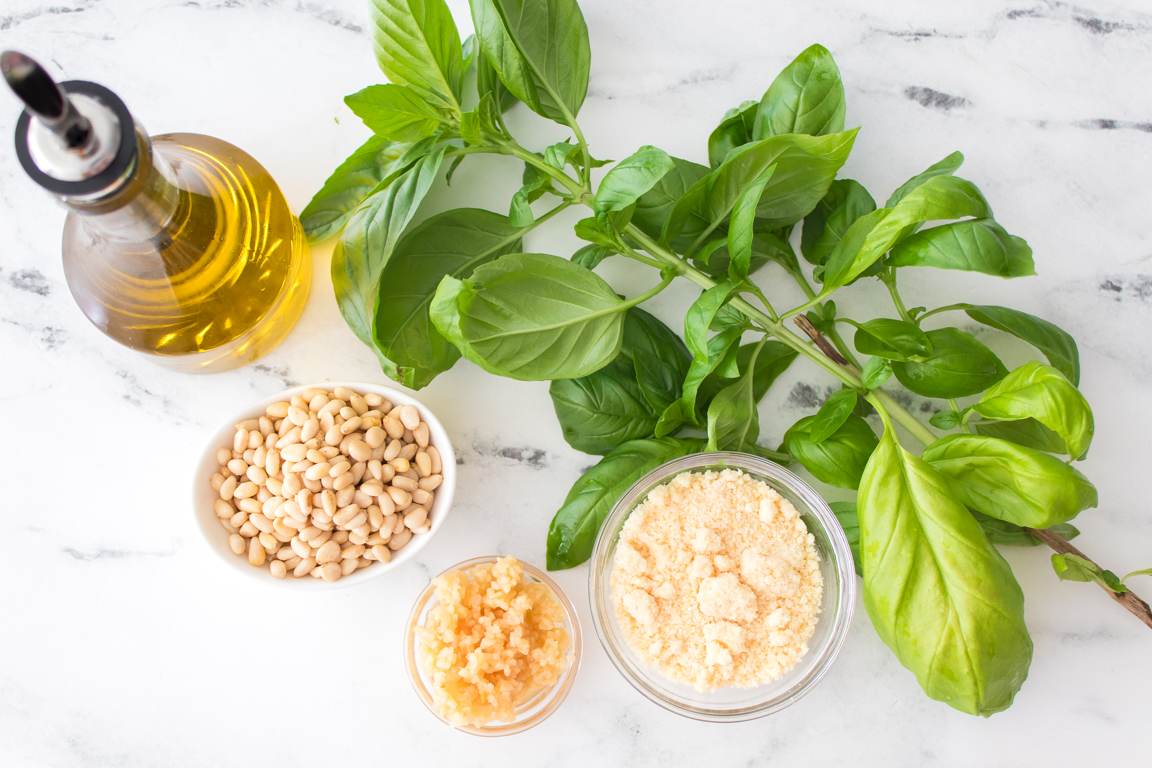 A marble surface with a bottle of olive oil, fresh basil, a bowl of pine nuts, grated parmesan, and minced garlic arranged neatly for making classic basil pesto.