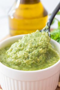 A spoon scoops classic basil pesto sauce from a white ramekin, with a bottle of olive oil in the background.