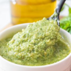 A spoon scoops classic basil pesto sauce from a white ramekin, with a bottle of olive oil in the background.