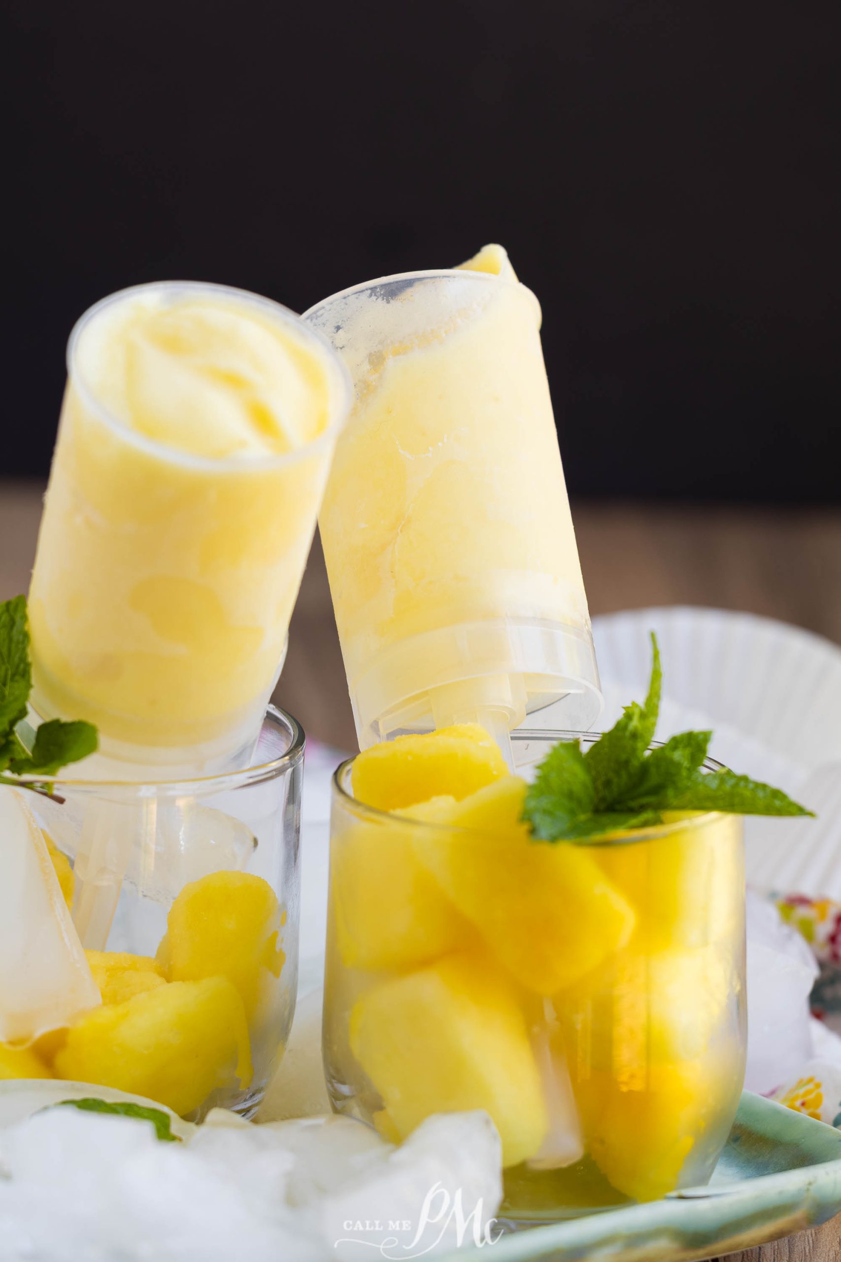 Two push-up pops with yellow frozen dessert are propped in glasses filled with pineapple chunks and garnished with fresh mint leaves.