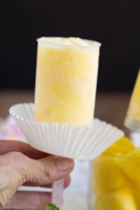 A hand holds a yellow frozen pop in a clear plastic mold, with a white paper liner underneath.
