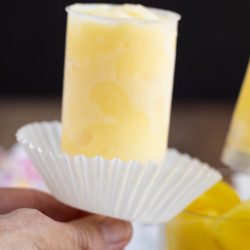 A hand holds a yellow frozen pop in a clear plastic mold, with a white paper liner underneath.