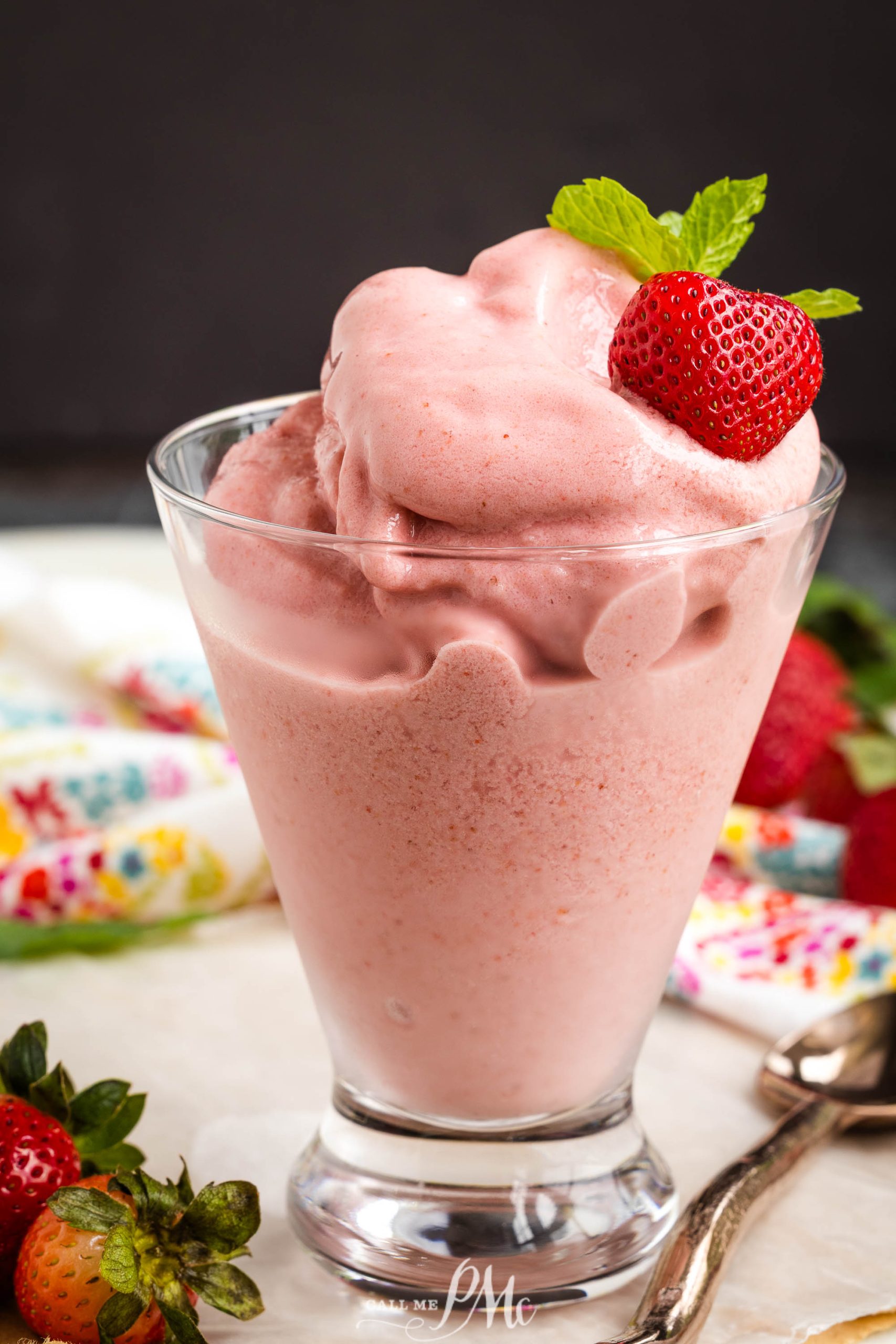 A glass filled with thick Boozy Strawberry Dole Whip smoothie, topped with a fresh strawberry and mint, sits on a table with strawberries and a spoon nearby.