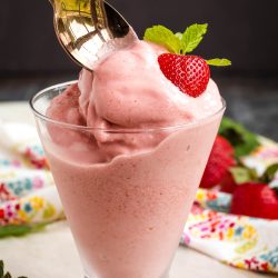 A spoon scoops pink Boozy Strawberry Dole Whip served in a glass, topped with a fresh strawberry and mint leaf.
