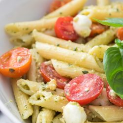 A bowl of Pesto Caprese Pasta Salad with penne pasta, vibrant pesto sauce, cherry tomato halves, small mozzarella balls, and a fresh basil leaf.