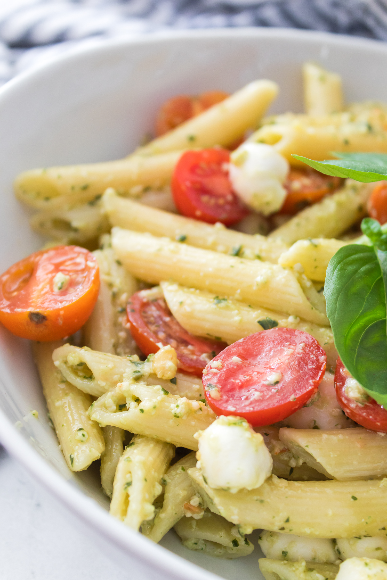A bowl of Pesto Caprese Pasta Salad with penne pasta, vibrant pesto sauce, cherry tomato halves, small mozzarella balls, and a fresh basil leaf.