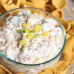 A glass bowl of Creamy Cowboy Caviar Dip with corn, peppers, and green onions, surrounded by crunchy corn chips on a speckled plate.