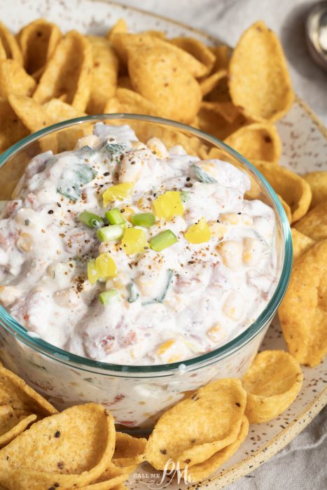 A glass bowl of Creamy Cowboy Caviar Dip with corn, peppers, and green onions, surrounded by crunchy corn chips on a speckled plate.