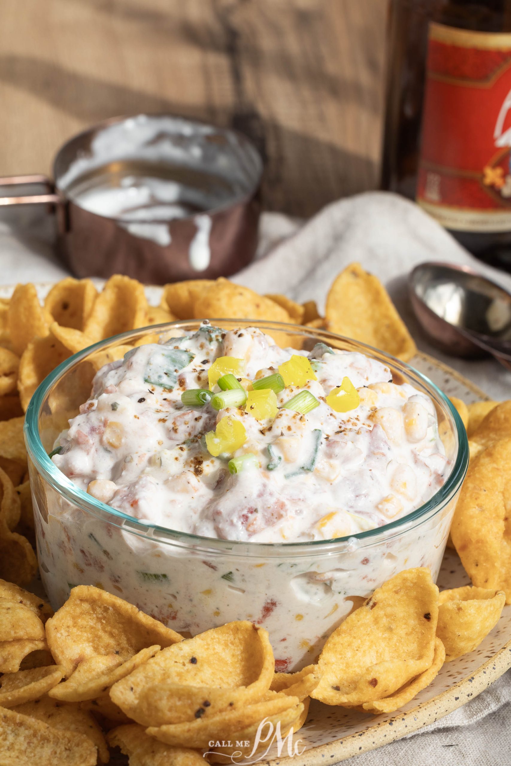 A glass bowl of appetizer garnished, surrounded by corn chips on a plate. A metal measuring cup with leftover dip is in the background.