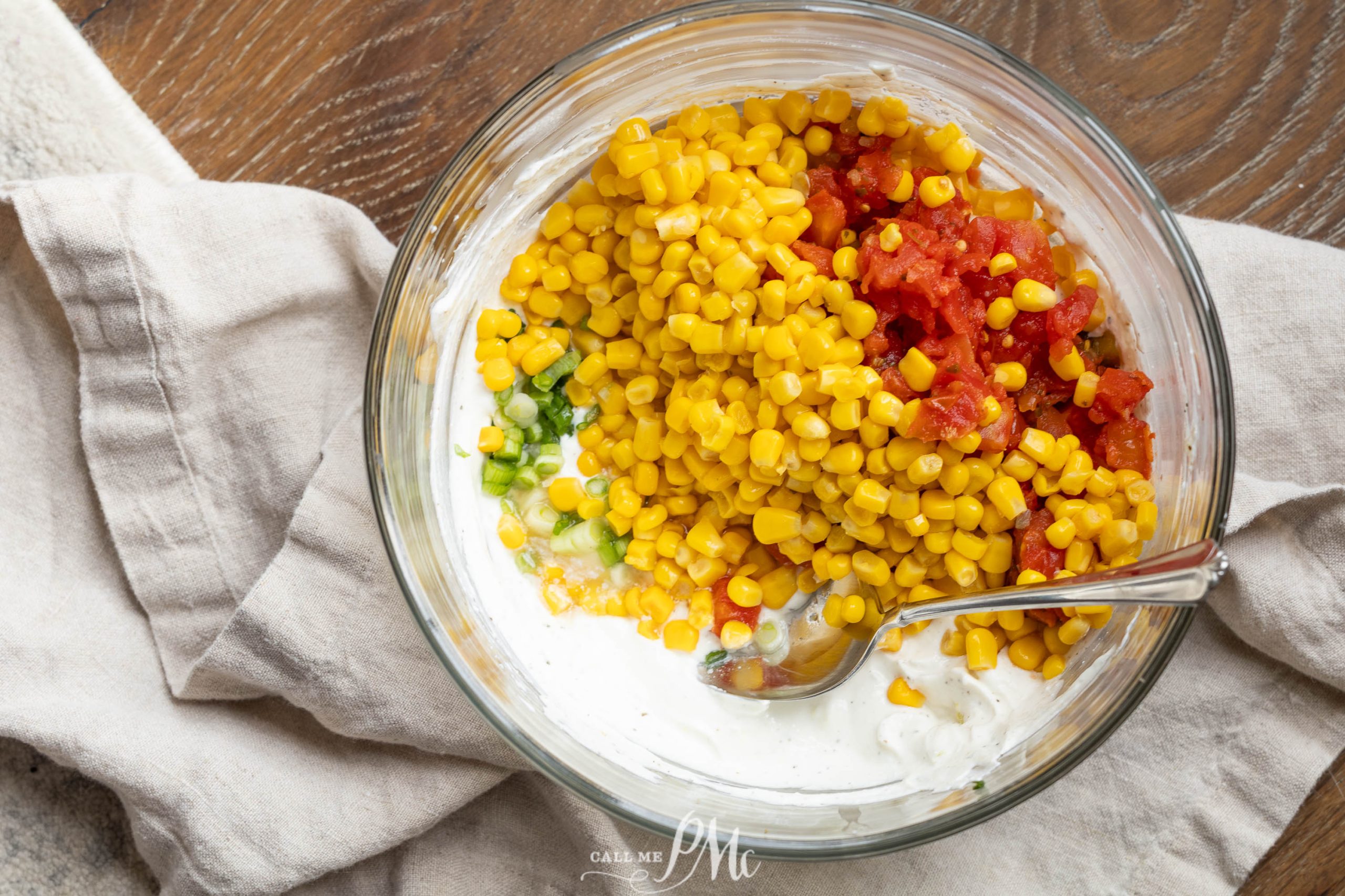 A glass bowl of Creamy Cowboy Caviar Dip with corn, diced tomatoes, chopped green onions, and creamy dressing sits with a spoon on a beige cloth and wooden surface.