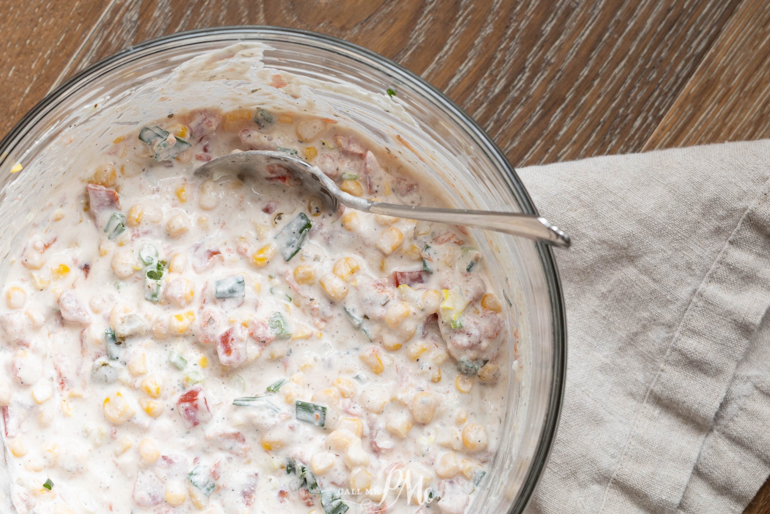 A glass bowl filled with Creamy Cowboy Caviar Dip, mixed with chopped vegetables and corn, with a silver spoon resting inside, placed on a wooden table next to a beige cloth napkin.