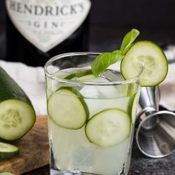A refreshing Cucumber Basil Gin Cooler, with ice, cucumber slices, and basil garnish, sits in front of a Hendrick's gin bottle and a metal jigger on a dark countertop.