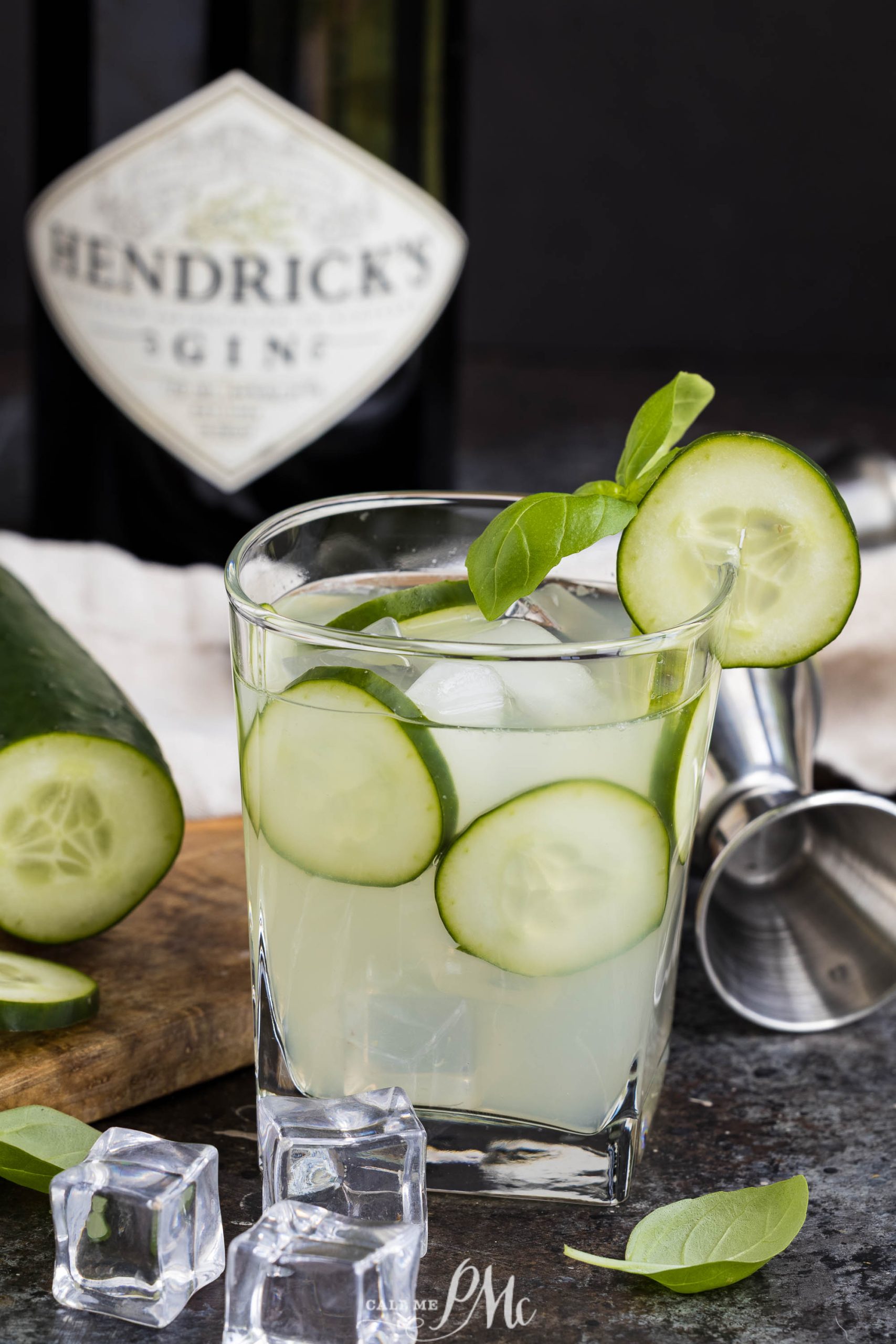 A refreshing Cucumber Basil Gin Cooler, with ice, cucumber slices, and basil garnish, sits in front of a Hendrick's gin bottle and a metal jigger on a dark countertop.