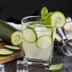 A glass of Cucumber Basil Gin Cooler with ice, garnished with cucumber slices and basil, sits on a counter next to a shaker, ice cubes, and a partially sliced cucumber.