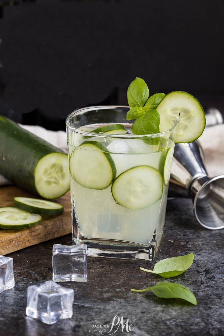 A glass of Cucumber Basil Gin Cooler with ice, garnished with cucumber slices and basil, sits on a counter next to a shaker, ice cubes, and a partially sliced cucumber.