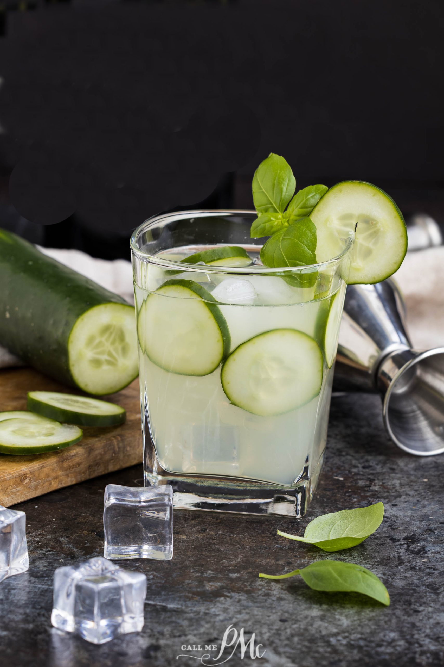 A glass of Cucumber Basil Gin Cooler with ice, garnished with cucumber slices and basil, sits on a counter next to a shaker, ice cubes, and a partially sliced cucumber.