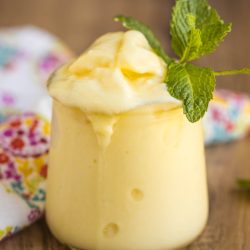 A glass of creamy yellow pineapple dessert topped with a sprig of fresh mint, placed on a wooden surface with a floral cloth in the background.