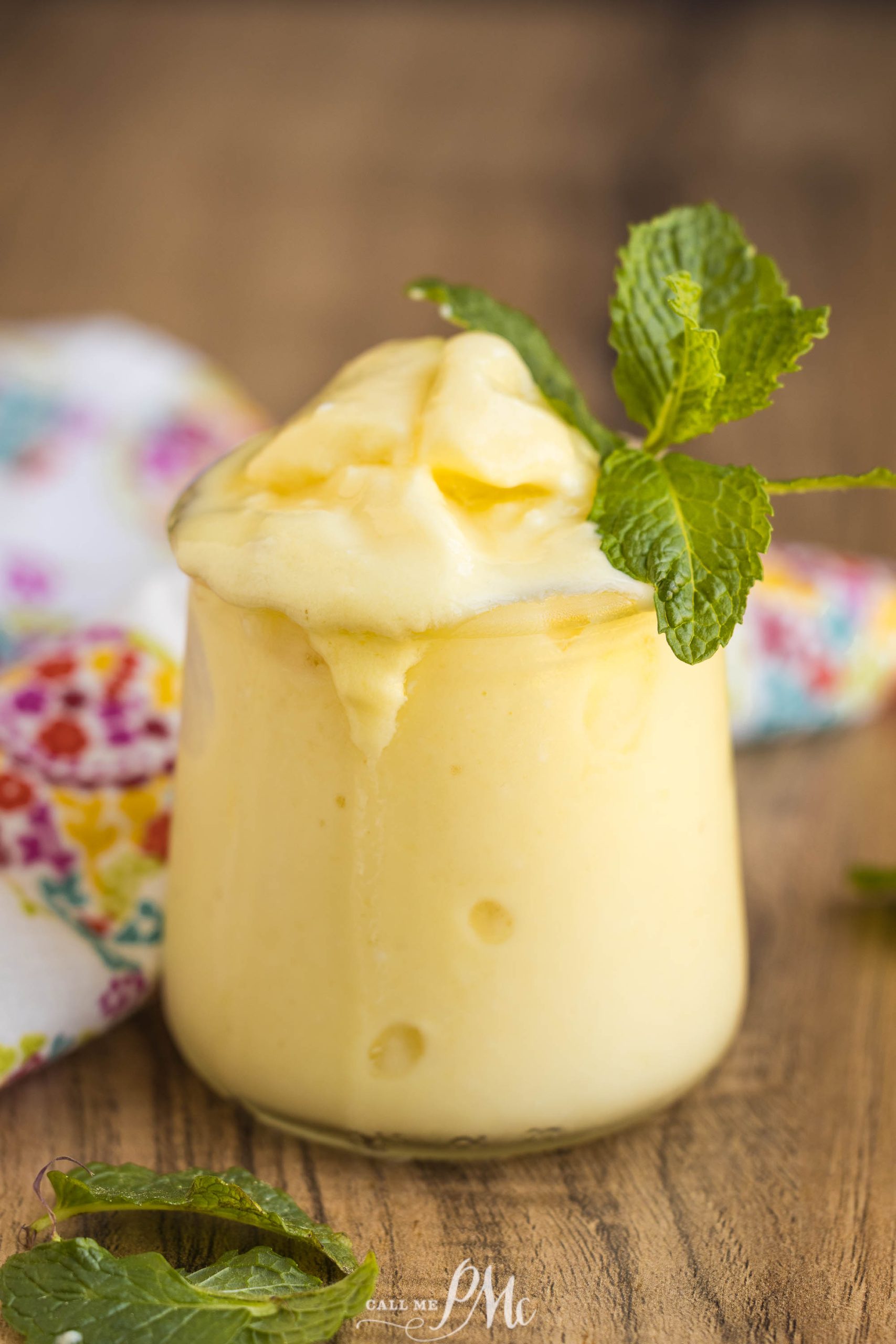 A glass of dairy-free boozy Dole Whip topped with a sprig of fresh mint, placed on a wooden surface with a floral cloth in the background.