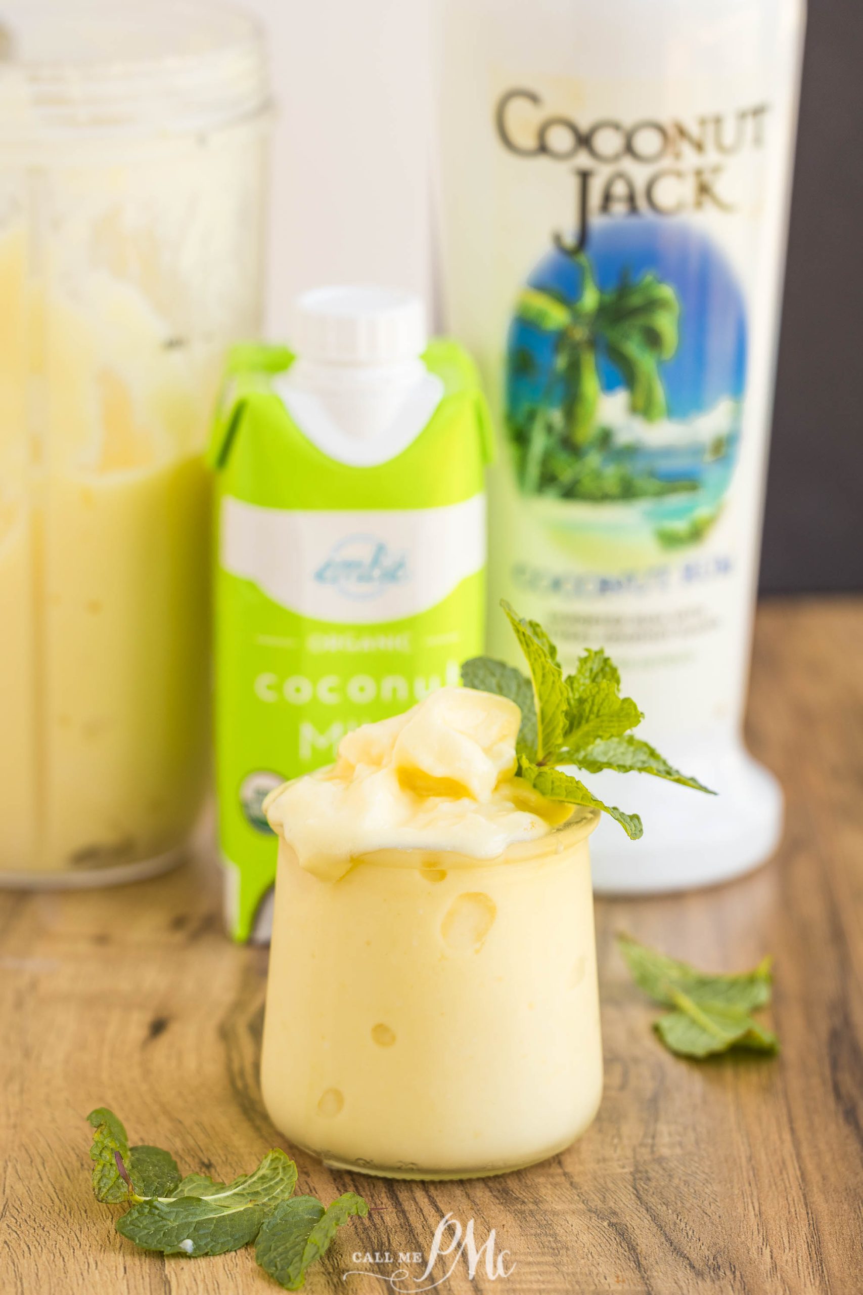 A creamy yellow drink garnished with mint sits on a wooden table, with coconut milk and coconut liqueur containers in the background.