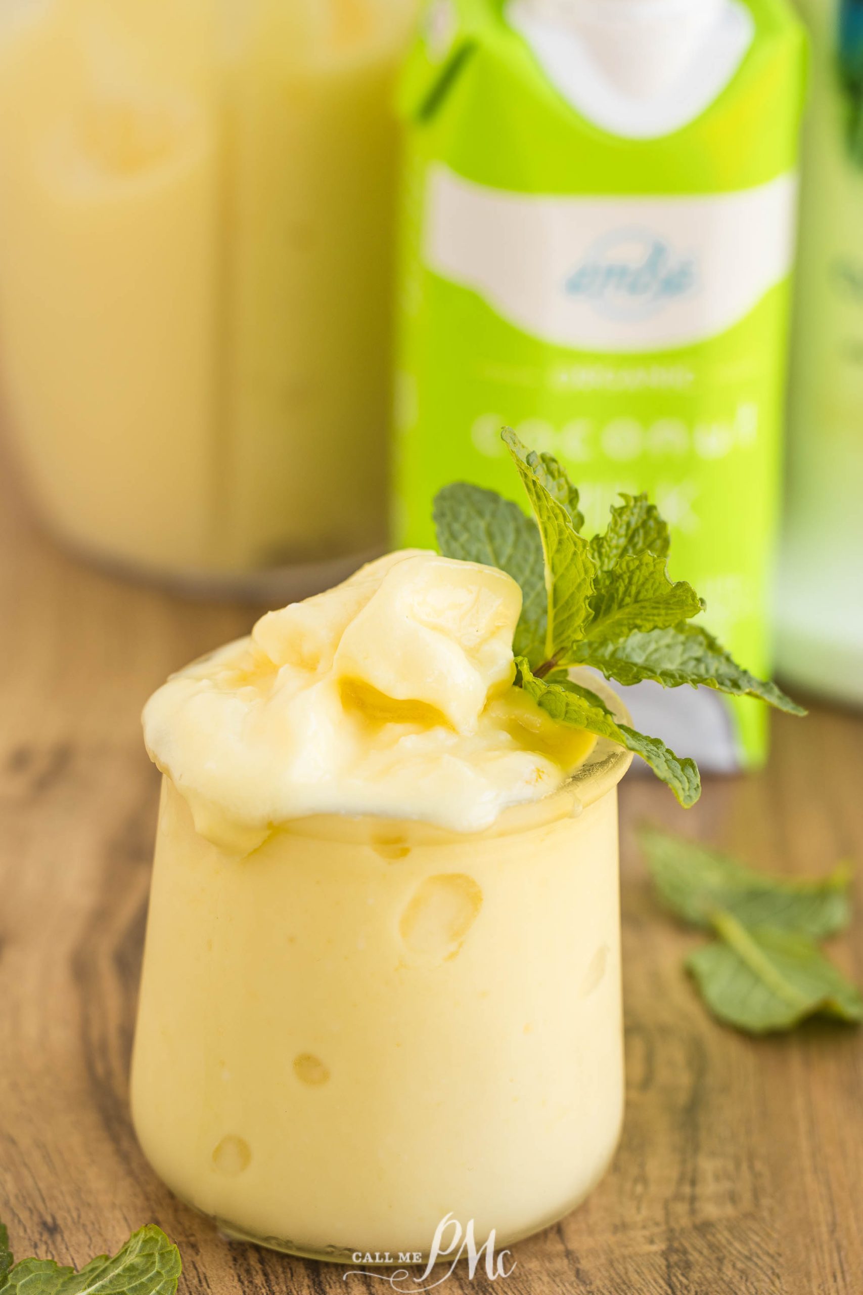 A small glass of dairy-free boozy Dole Whip smoothie garnished with a mint sprig, set on a wooden surface with fresh mint leaves and green drink cartons in the background.