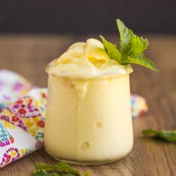 A glass of creamy yellow pineapple Dole Whip topped with a sprig of fresh mint, placed on a wooden surface with a colorful cloth nearby.