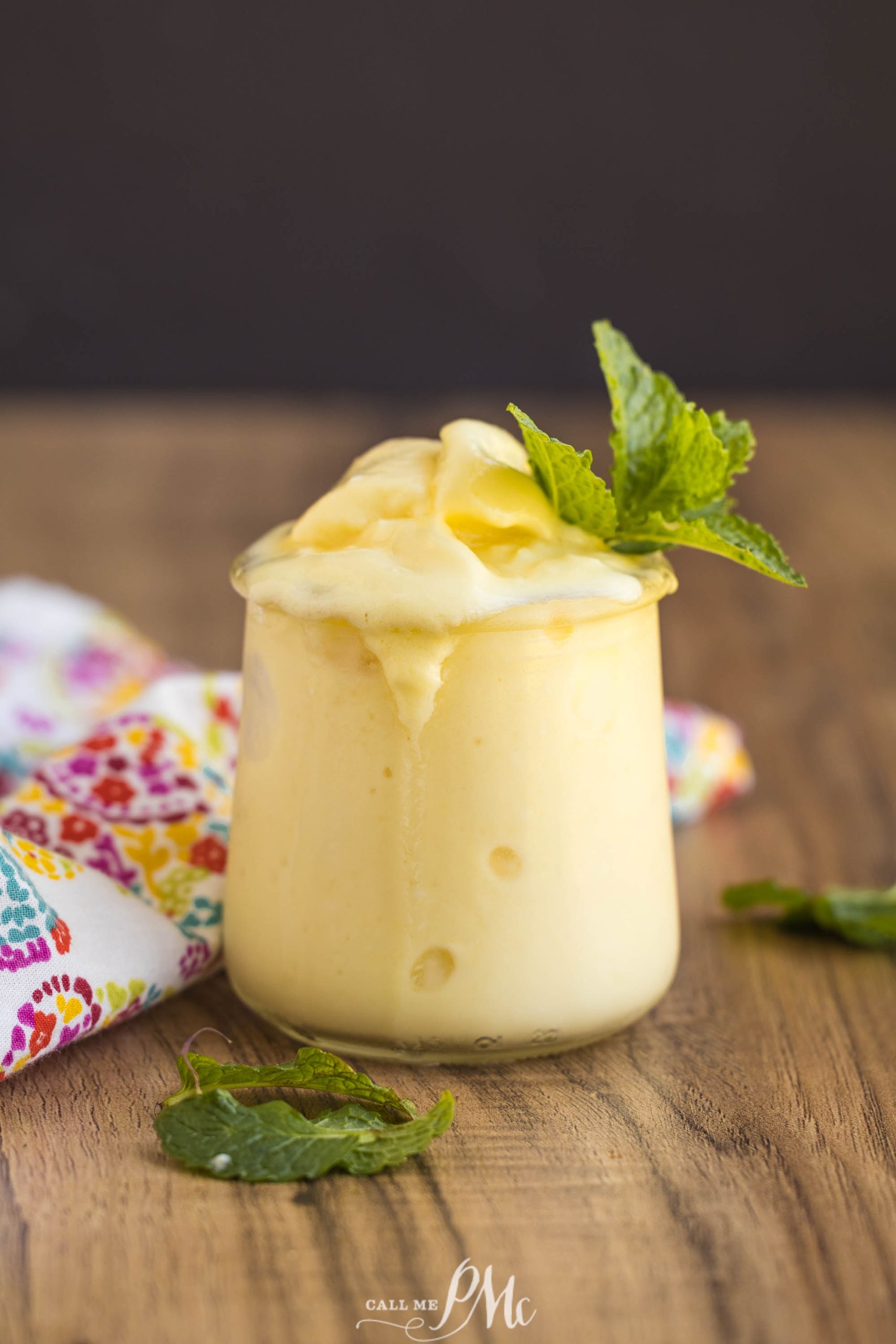 A glass of creamy yellow pineapple Dole Whip topped with a sprig of fresh mint, placed on a wooden surface with a colorful cloth nearby.
