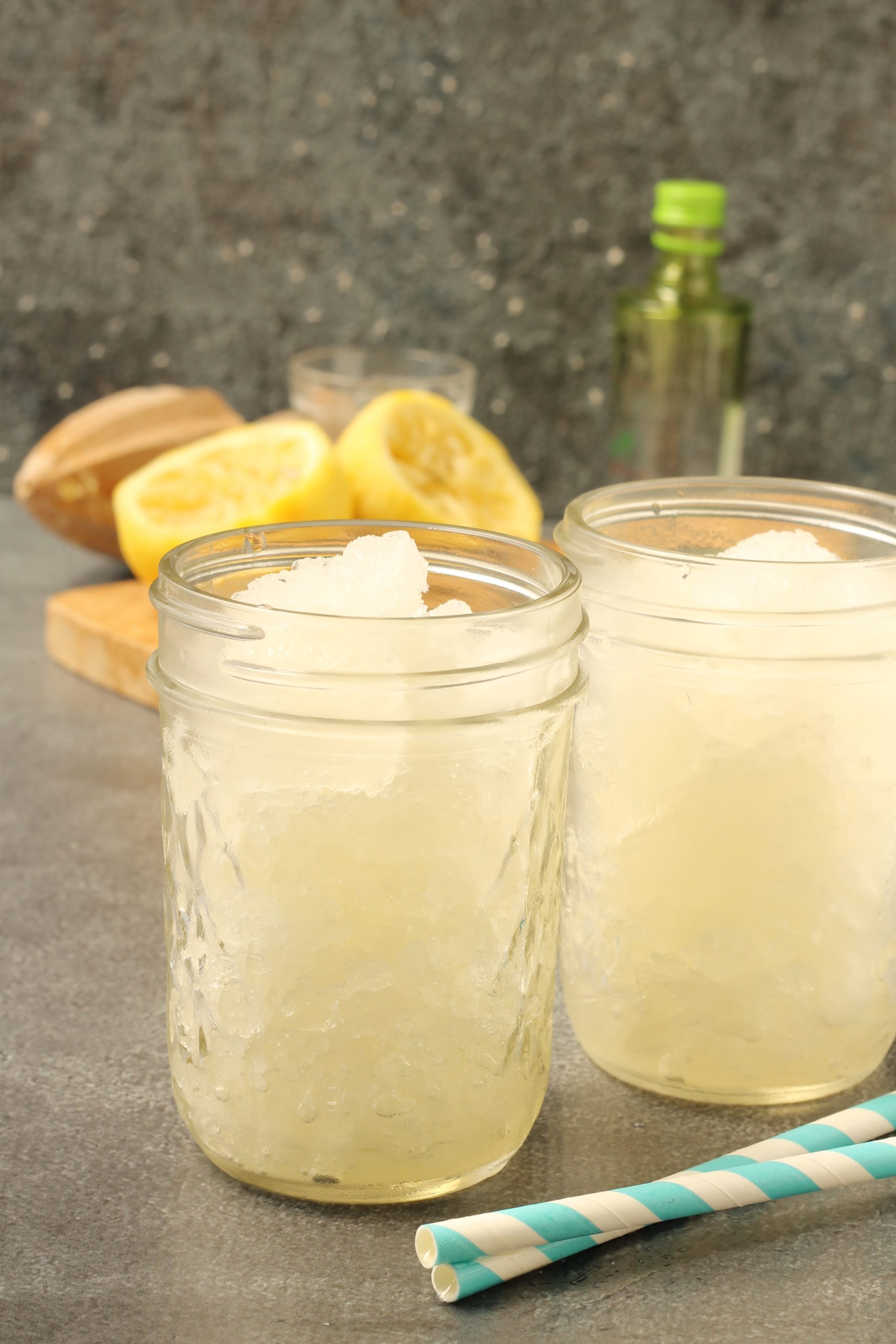 Two mason jars filled with Frozen Lemonade Gin Cocktail shaved ice sit on a tabletop, with lemon halves, a bottle, and two striped straws nearby.