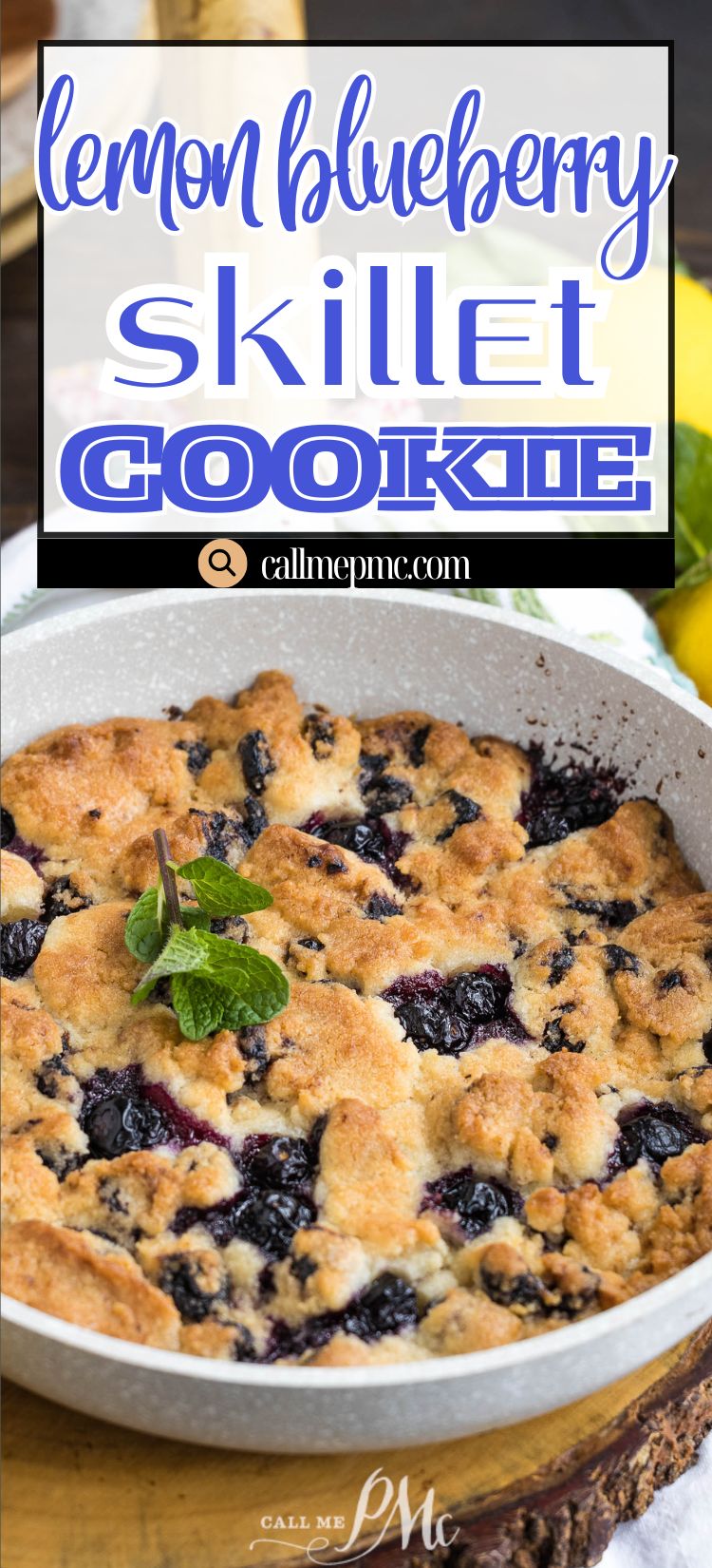 A baked lemon blueberry skillet cookie garnished with fresh mint, displayed in a round pan on a wooden surface. A lemon and a cloth are visible in the background.