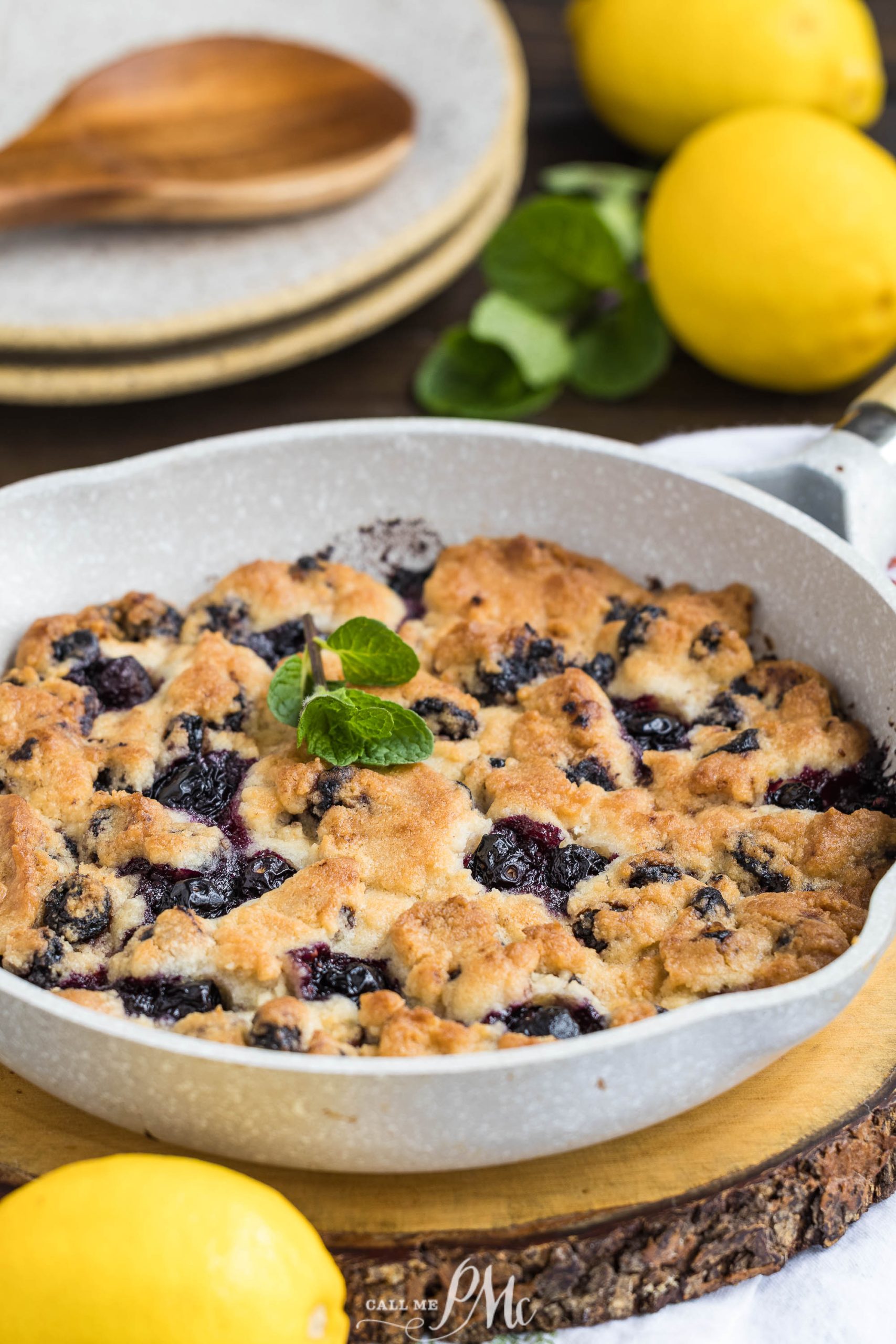 A baked blueberry cobbler in a round dish, garnished with a mint sprig, surrounded by fresh lemons and wooden plates in the background.