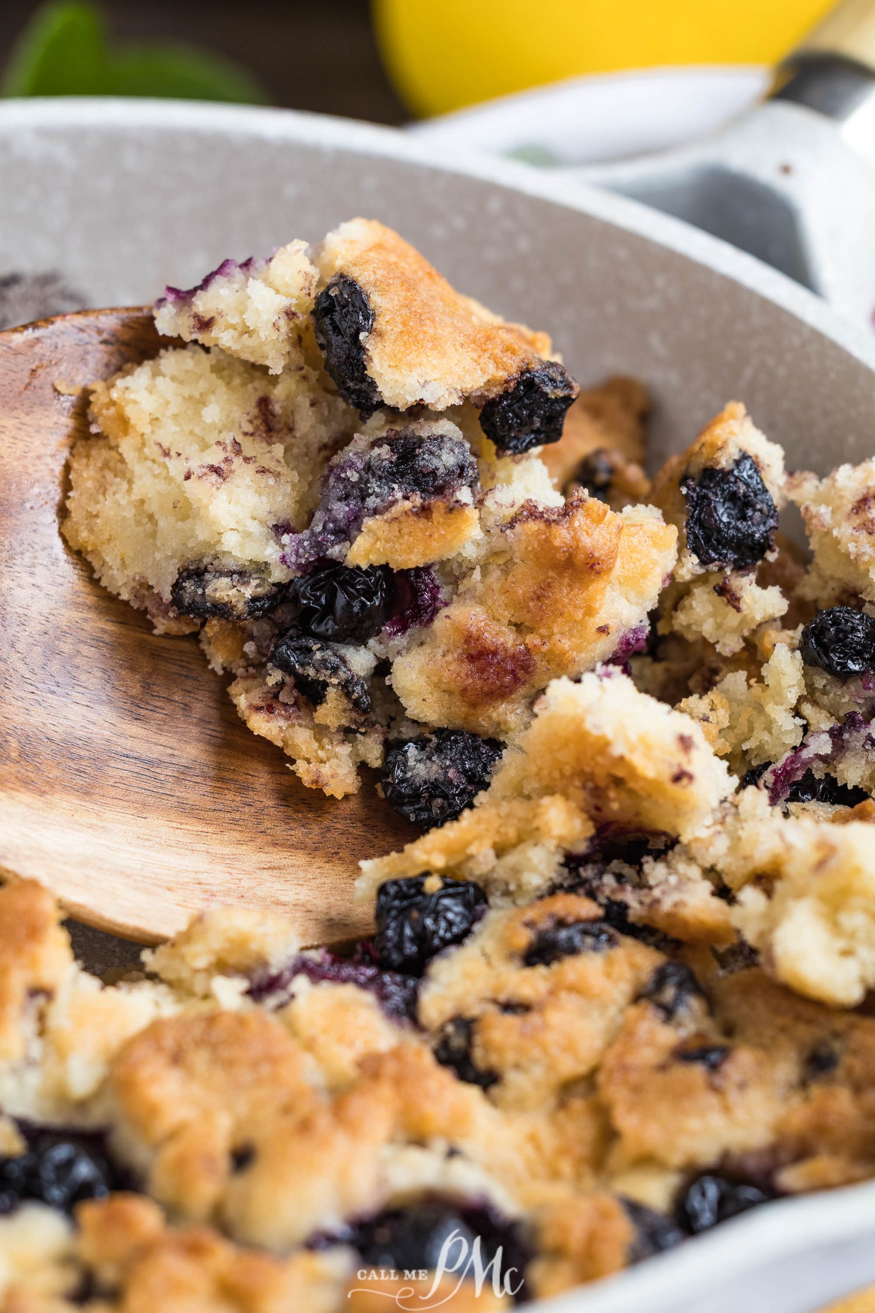 A close-up of a wooden spoon scooping out a portion of blueberry cobbler from a pan, showing golden crust and baked blueberries.