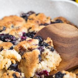 A wooden spoon scoops out a portion of blueberry cobbler from a white dish, showing golden-brown biscuit topping and cooked blueberries.
